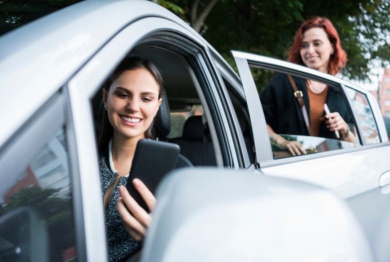 duas mulheres sorrindo segurando o celular nas mãos, uma no volante e a outra entrando atrás como passageira